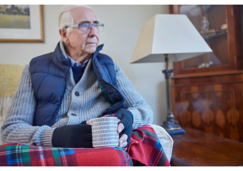 Elderly Man sat in a chair, looking out of the window with a blanket and gloves on and holding a hot drink