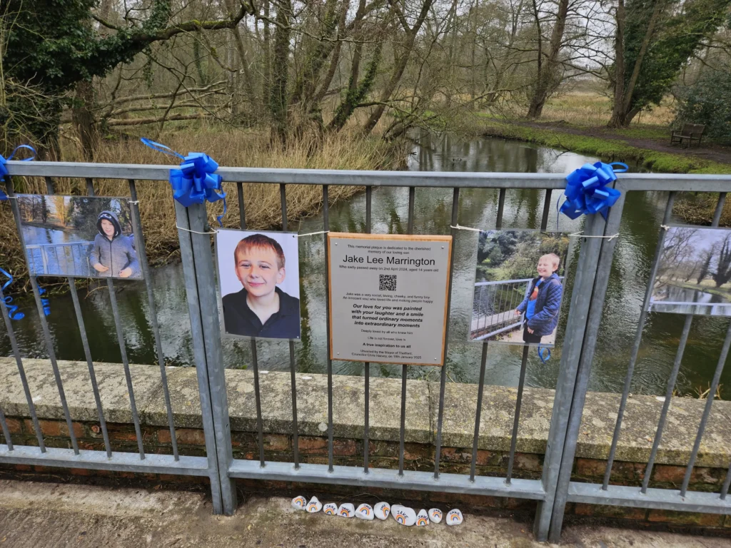 A memorial display on a metal bridge railing, dedicated to Jake Lee Marrington. The display features three photos of Jake at different ages, along with a plaque in the center containing a tribute message. Blue ribbons are tied to the railings, and small painted stones are placed at the base. The background shows a peaceful, wooded area with a calm river flowing beneath the bridge.