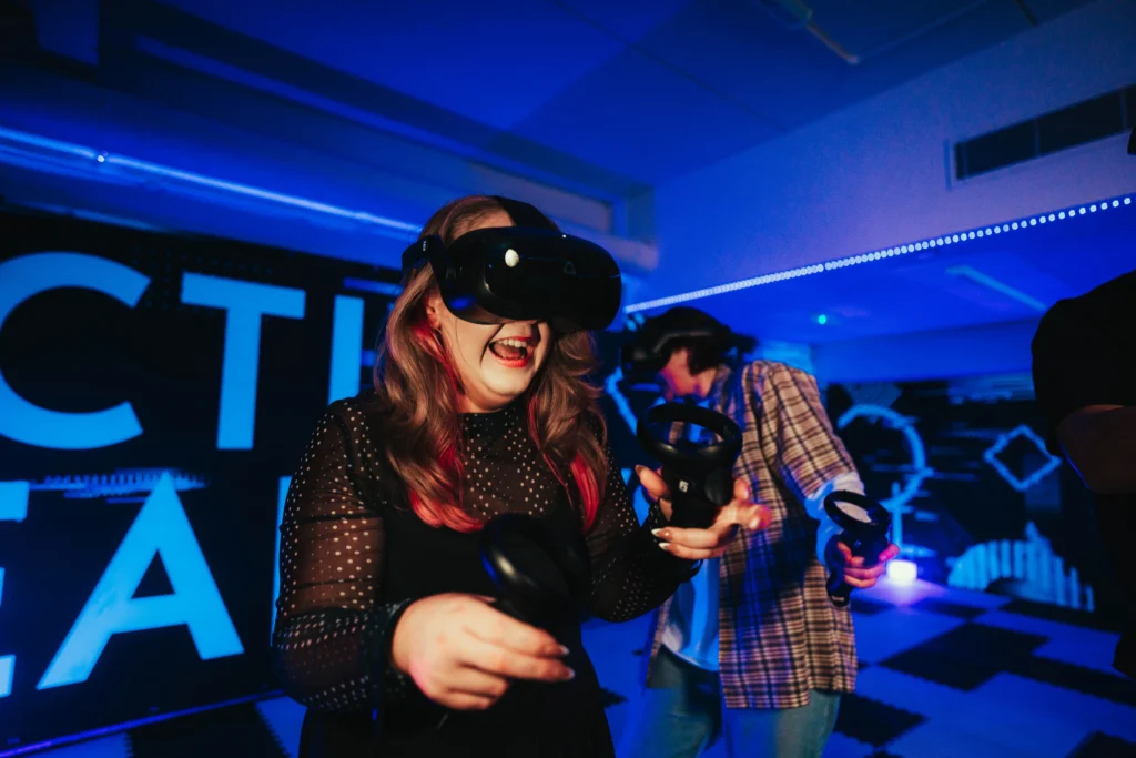 A woman with long hair and pink streaks laughs while immersed in a VR gaming session. She wears a black dress with sheer polka-dot sleeves and holds two VR motion controllers. Behind her, other players in VR headsets interact with the virtual environment. The setting is a neon-lit gaming space with a futuristic atmosphere.