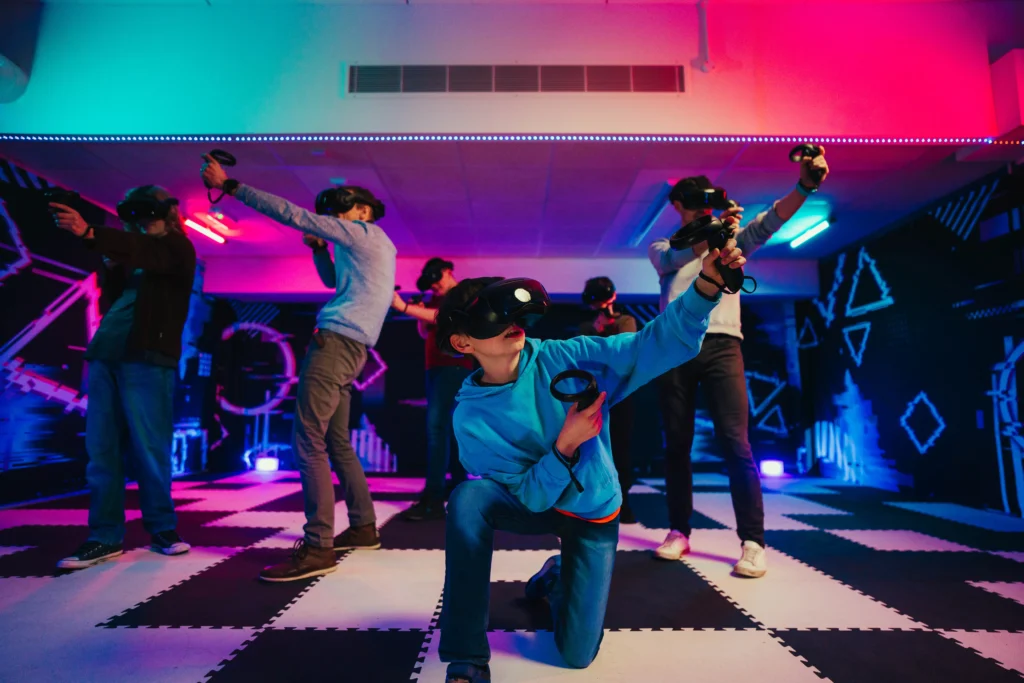 A group of people wearing VR headsets and holding motion controllers engage in a virtual reality gaming session in a futuristic, neon-lit room. A child in a blue hoodie is kneeling in the foreground, aiming their controller, while others stand in action poses behind them. The room features a checkered floor and glowing geometric wall designs, illuminated by blue and pink lights.