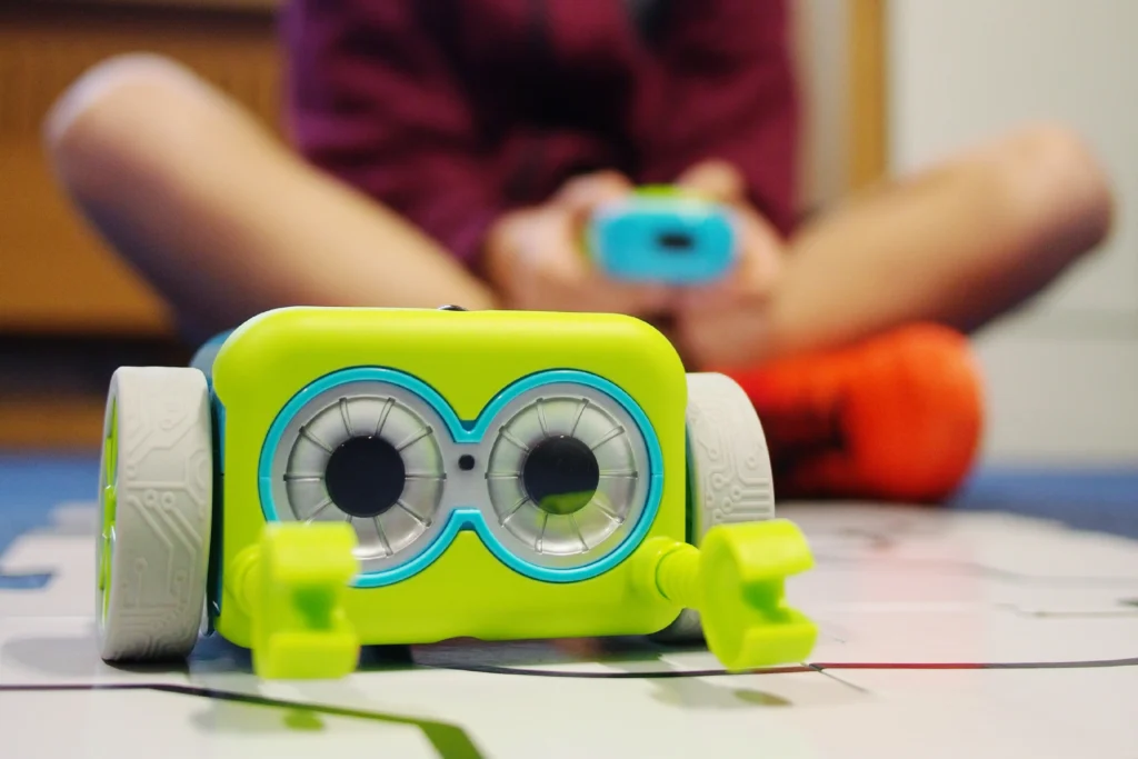 A bright green and blue toy robot with large, round "eyes" and small mechanical arms sits on a play mat with a track design. In the blurred background, a child wearing a maroon top and orange sneakers is sitting cross-legged, holding a blue remote control and controlling the robot. The setting appears to be an interactive learning or play session with robotics.
