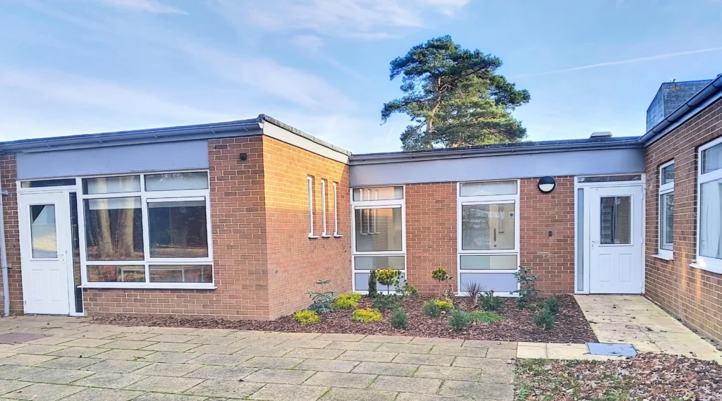 The front view of a single-story school building made of red brick with large windows and a white entrance door. A small landscaped garden with young plants and shrubs is in the foreground, with a clear blue sky above.