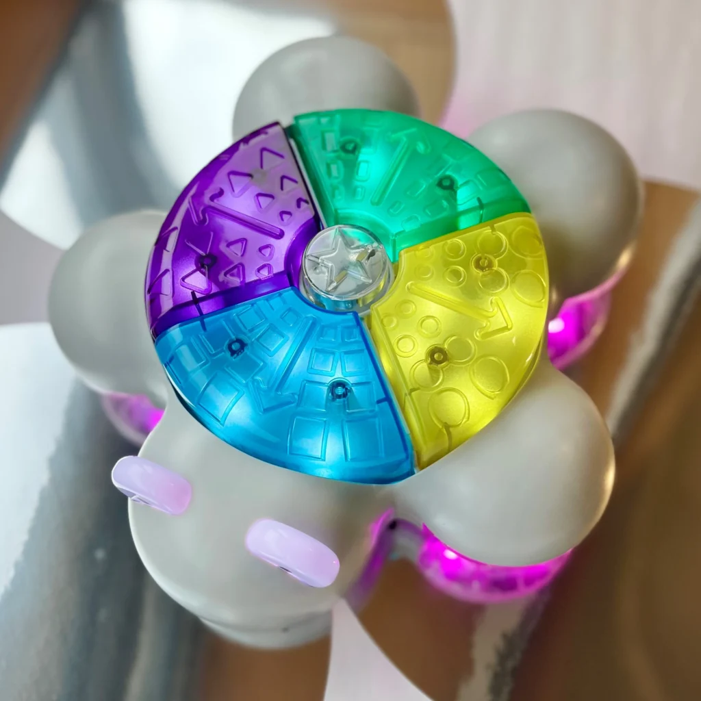 A close-up of a glowing robotic toy with a round body and translucent, colorful panels in purple, blue, green, and yellow. The toy emits a soft pink and purple light from its base, giving it a futuristic appearance. The reflective surface beneath the toy enhances its illuminated effect. It is an interactive or educational tool.