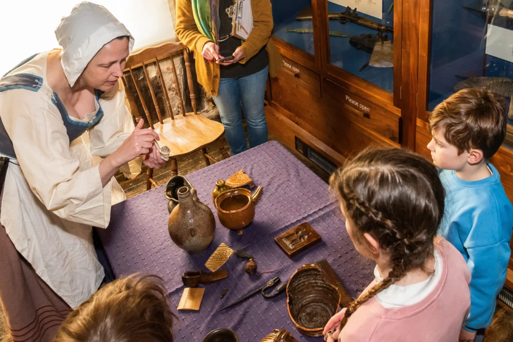 A costumed museum educator dressed in a Tudor-style outfit, including a white bonnet and historical dress, engages a group of young children in a hands-on learning session. She is demonstrating an artifact while the children, dressed in modern clothing, watch attentively. A table covered with a purple cloth displays various historical objects such as pottery, wooden combs, and metal tools. Behind them, a museum display case holds additional artifacts.