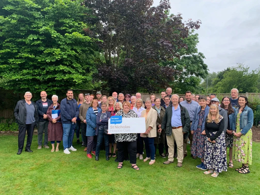 A large group of Thetford Open Gardens volunteers and supporters gathers outdoors on a lush green lawn, posing for a group photo in front of trees and a wooden fence. Many are dressed in smart casual attire, with some holding folders or wearing jackets. In the center, a woman holds a sign that reads "St Nicholas Hospice Care – because you matter." The group is celebrating their efforts in supporting the hospice through their open gardens initiative. Everyone is smiling, creating a warm and welcoming atmosphere.