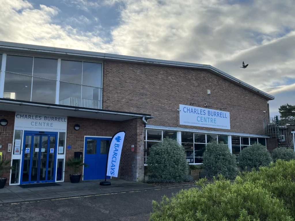 An exterior view of the Charles Burrell Centre, a large brick building with blue-framed windows and doors. Signs above the entrance and on the side of the building read "CHARLES BURRELL CENTRE" with the tagline "Uniting Community, Learning & Business Under One Roof." A Barclays-branded flag stands near the entrance, and neatly trimmed bushes line the front of the building. The sky is partly cloudy, and a bird is seen flying above the building.