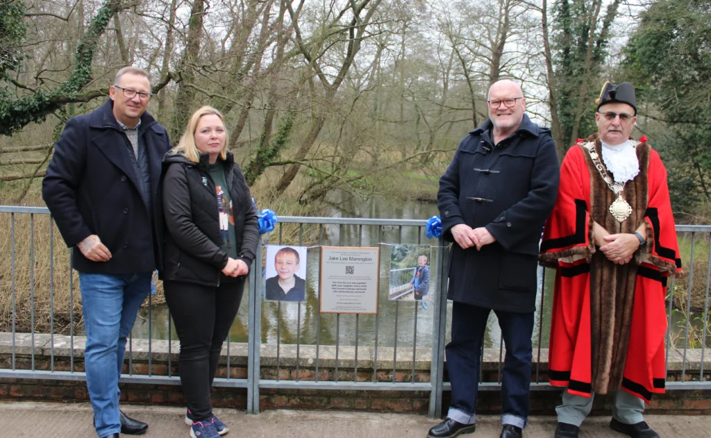 Four people stand in front of a memorial plaque dedicated to Jake Lee Marrington on a metal bridge railing. The group includes two men in dark coats, a woman in a black jacket, and a man in ceremonial attire wearing a red robe and chain of office. Photos of Jake and blue ribbons are attached to the railing behind them. The background shows a wooded area with a river running beneath the bridge.