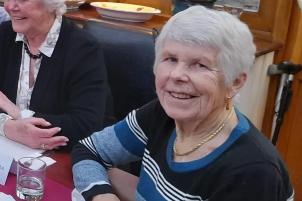 Liz Hodgson, an elderly woman with short white hair, smiles warmly while seated at a dining table. She is wearing a striped black, blue, and white sweater along with a gold necklace. In the background, another woman in a black cardigan and white blouse is engaged in conversation. A glass of water, a name card, and a plate with food remnants are visible on the table, suggesting a social gathering. A walking aid is positioned nearby.