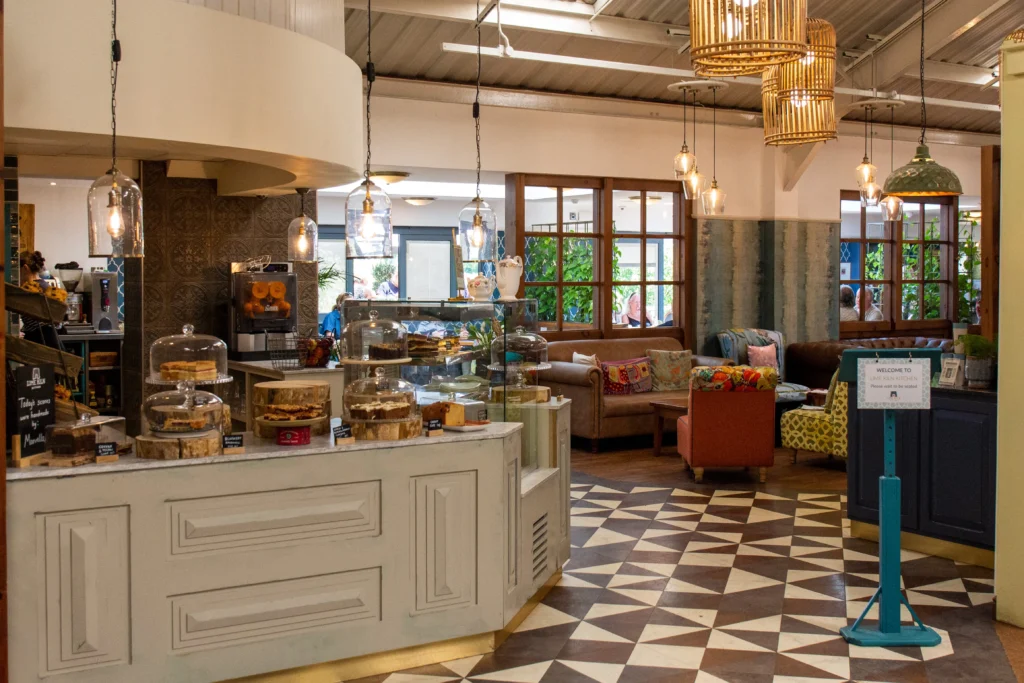 A cozy café interior featuring a counter displaying a variety of cakes under glass domes. The space includes comfortable seating with colorful cushions, wooden furniture, and patterned floor tiles. Warm lighting from hanging lamps adds to the inviting ambiance.