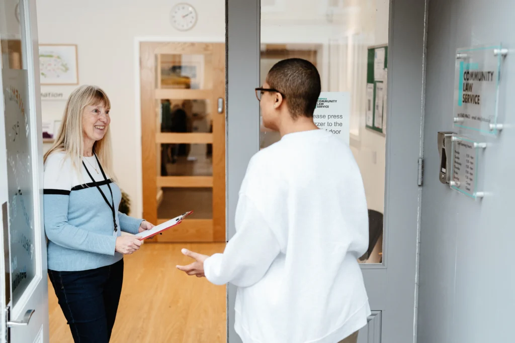 A smiling receptionist with long blonde hair, wearing a light blue and white sweater, greets a visitor at the entrance of the Norfolk Community Law Service office. The visitor, wearing glasses and a white jumper, gestures while speaking. A sign on the door reads "Community Law Service."