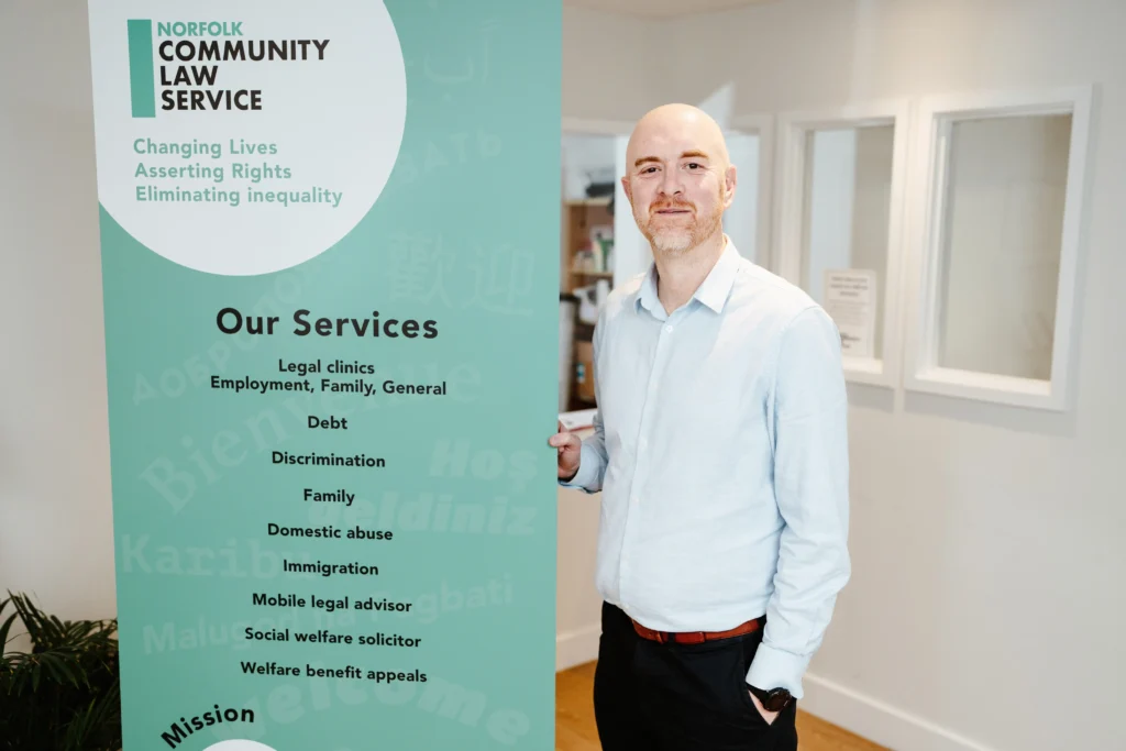 A bald man in a light blue shirt stands next to a large green Norfolk Community Law Service banner listing services such as legal clinics, debt advice, domestic abuse support, and welfare benefit appeals. The banner includes the organization's mission to "change lives, assert rights, and eliminate inequality."