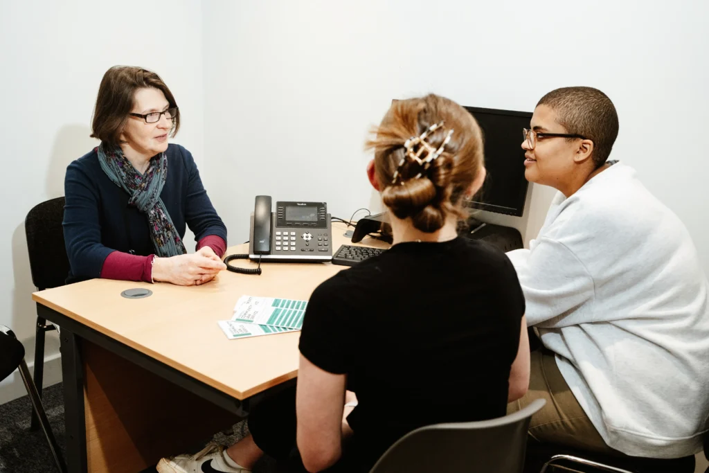 A consultation room where a woman with short brown hair, wearing glasses and a dark scarf, sits across from two young clients. One is wearing a white hoodie, the other a black t-shirt. A desk with a telephone, computer monitor, and information leaflets is in front of them as they discuss legal matters.