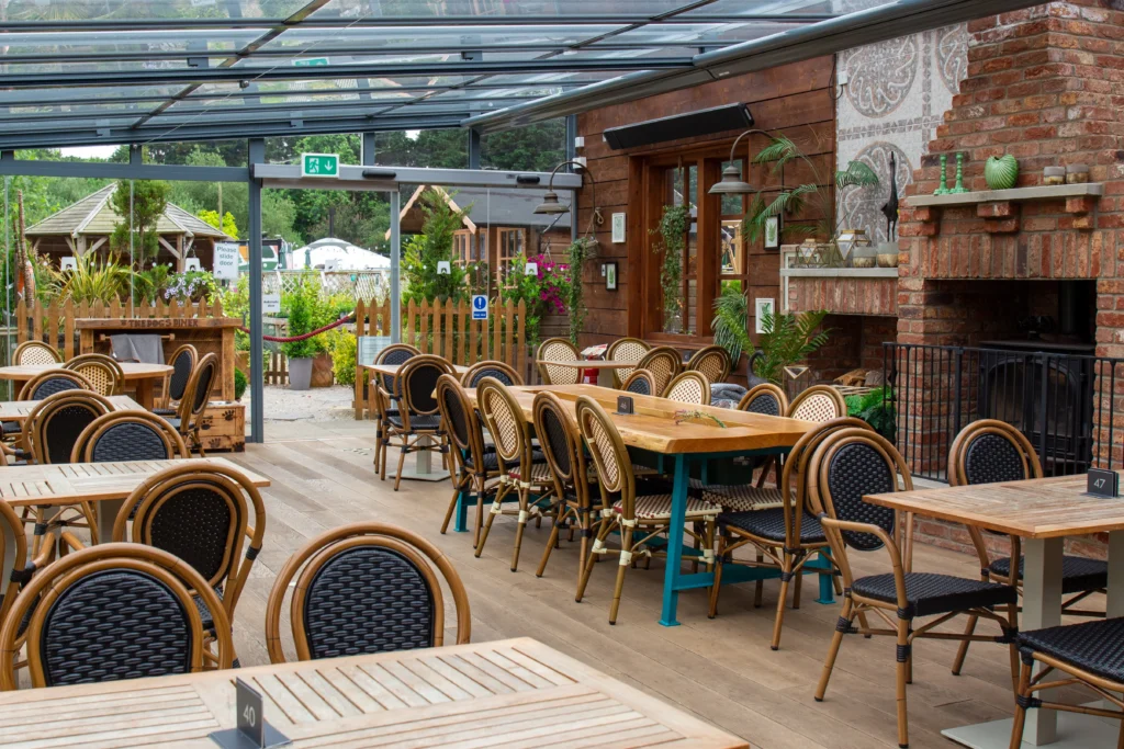 A bright, airy dining area with wooden tables and wicker chairs arranged neatly under a glass roof. The space is decorated with potted plants and greenery, creating a fresh and inviting atmosphere. Outside the glass walls, there’s a garden area with more greenery and wooden structures.