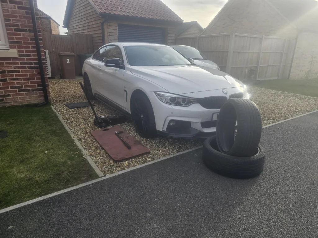 A white BMW parked on a gravel driveway with a jack lifting one side of the car. A mat with tools is placed nearby, and two tires are stacked on the asphalt next to the car. The background features brick houses, wooden fences, and bins marked with the number 2. Sunlight streams in from the right side of the image.