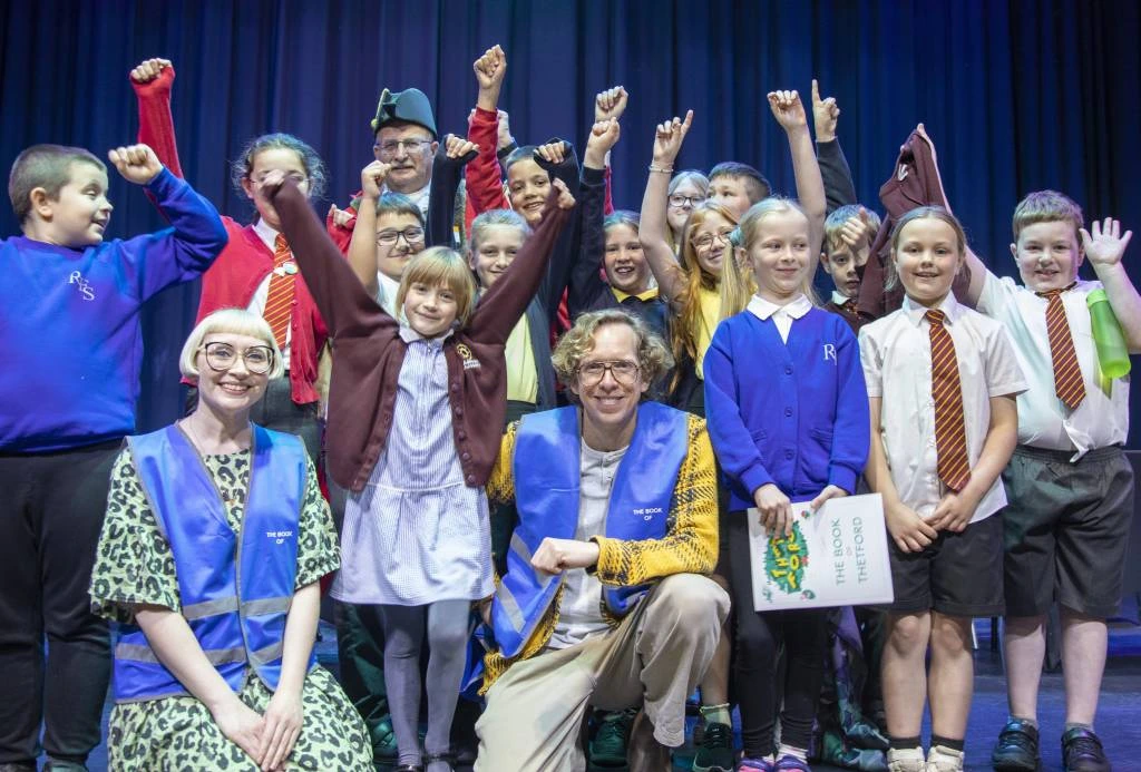 A large group of children in school uniforms and two adults in blue vests celebrate on a stage, raising their arms in the air. A man kneels at the front, smiling, while a young girl in a brown cardigan stretches her arms wide. Behind them, a man in a tricorn hat and red coat joins in the cheer. A blue curtain serves as the backdrop, and one child holds a booklet titled "The Book of Thetford."