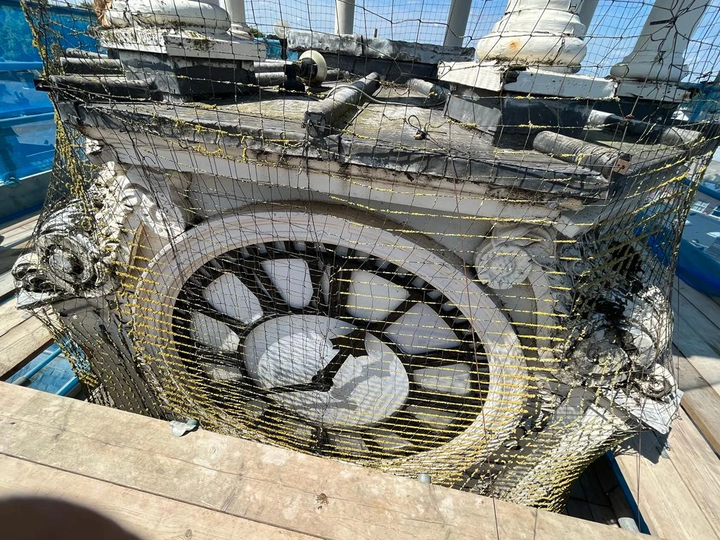 The exterior of an old clock tower undergoing restoration, with scaffolding and protective netting surrounding the structure. The ornate white clock face is weathered, with black hands positioned in the center. The surrounding architectural details, including decorative moldings and metal elements, show signs of wear and exposure to the elements.