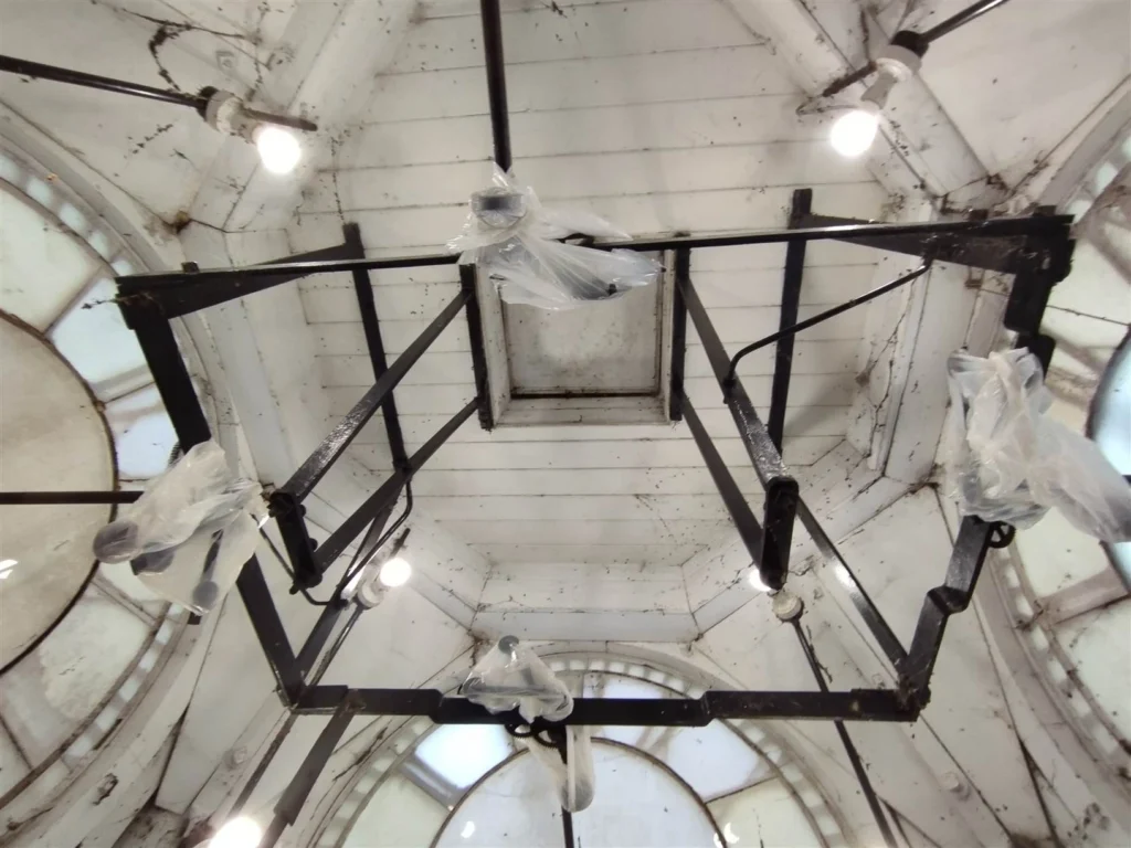 A view from inside the clock tower, looking up at the aged wooden ceiling with black metal framework supporting the clock mechanism. Several light bulbs illuminate the space, while plastic coverings are wrapped around some components. The surrounding clock faces, partially visible, allow natural light to filter through.