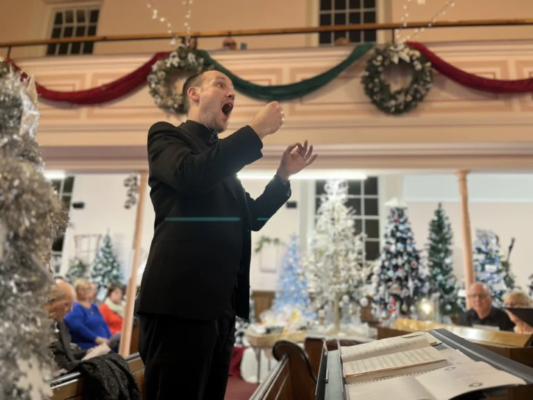 A man in a black suit conducts a choir in a festively decorated venue. He is mid-gesture with an expressive face, and the background features an audience, Christmas trees, and wreaths with red and green garlands.