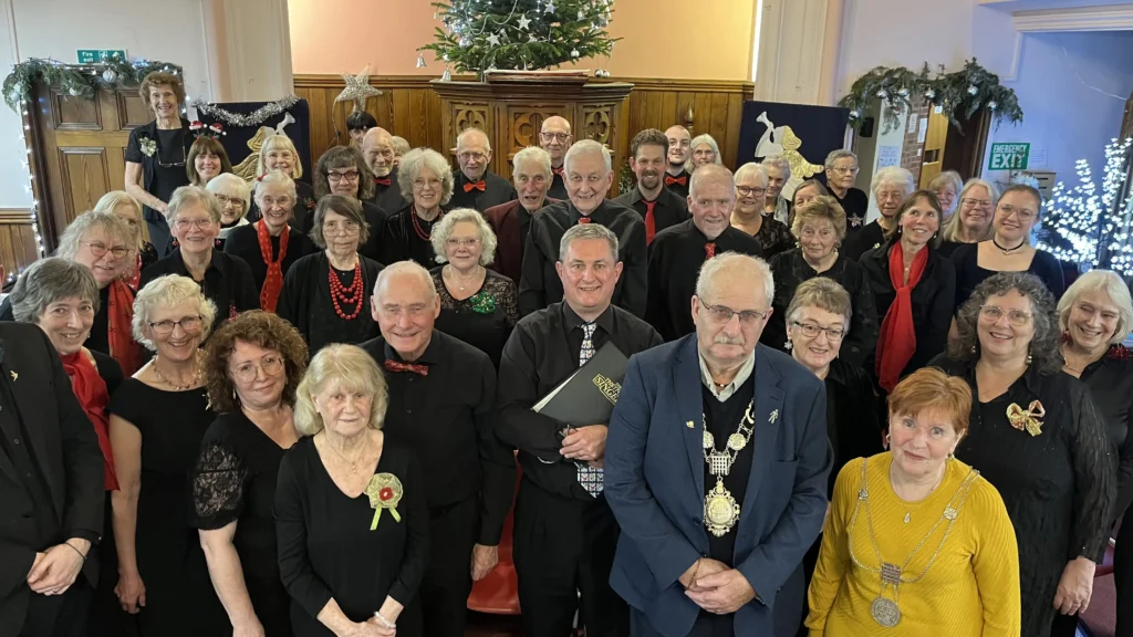 A group of men and women dressed in black with red and green accents stand together in a hall decorated for Christmas. Some hold choir folders, while others smile. The mayor, wearing ceremonial chains, stands at the front.