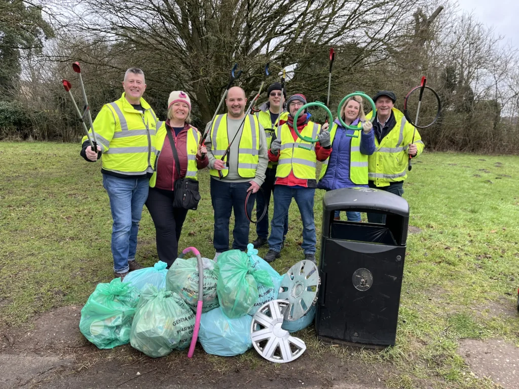 A group of eight volunteers wearing high-visibility vests pose in a grassy area with litter-picking tools. They smile and raise their equipment triumphantly. In front of them, several full green bags of collected rubbish, a discarded hubcap, and a pink plastic tube are piled next to a black bin. The background features leafless trees and a cloudy sky.