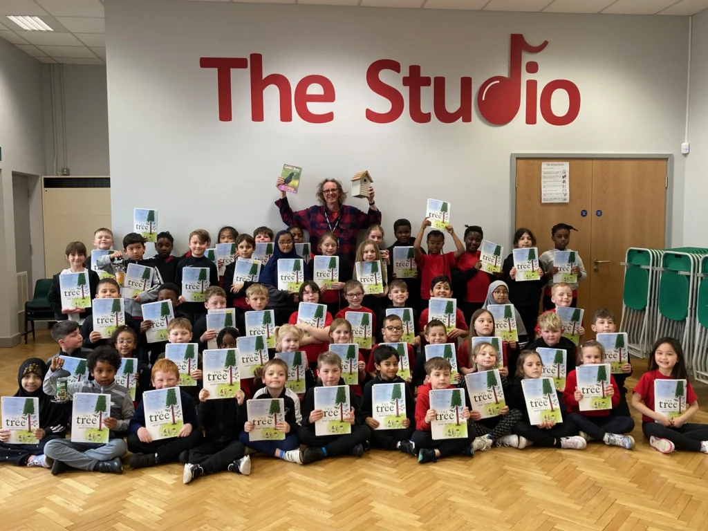 A large group of schoolchildren pose in a school hall beneath a red sign that reads “The Studio.” The children are seated and standing in rows, each holding a book titled "The Tree." An adult stands behind them, smiling and holding up a copy of the same book and a small wooden birdhouse. The children wear a mix of red, black, and grey clothing. A stack of green plastic chairs is visible to the right.