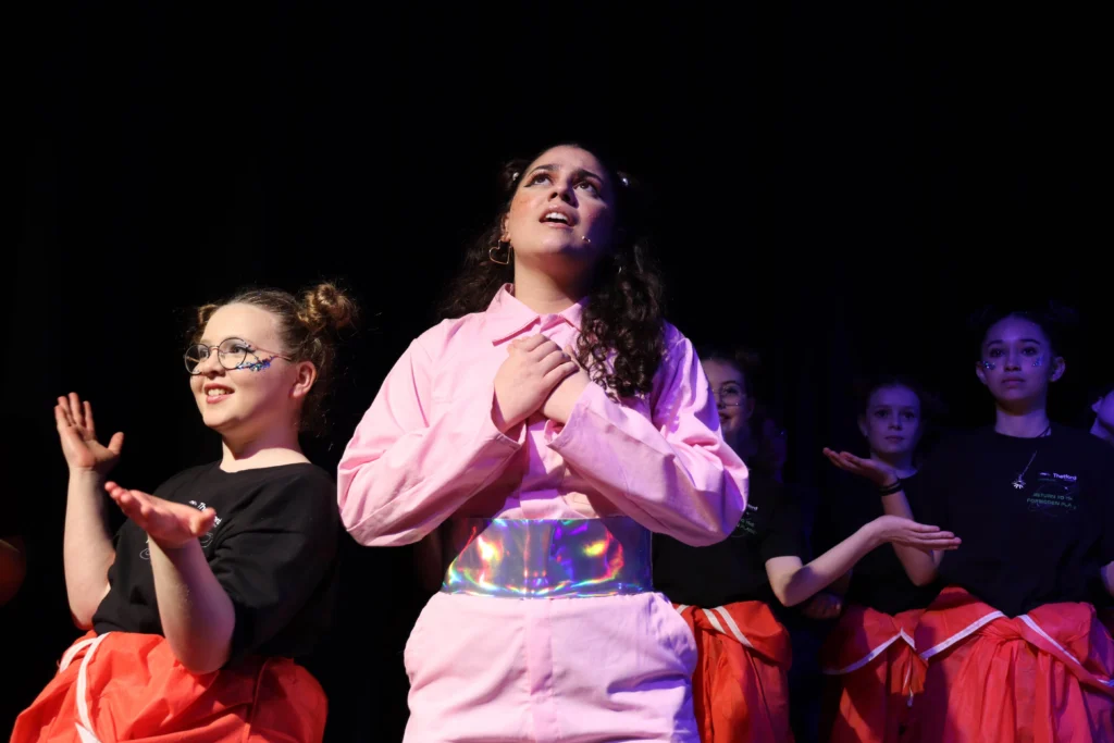 A theatrical performance on stage featuring a young woman in a pink jumpsuit with an iridescent belt, hands clasped to her chest, gazing upwards with an emotional expression. Behind her, several performers dressed in black shirts and red skirts with decorative makeup and accessories engage in expressive hand gestures under dramatic stage lighting.