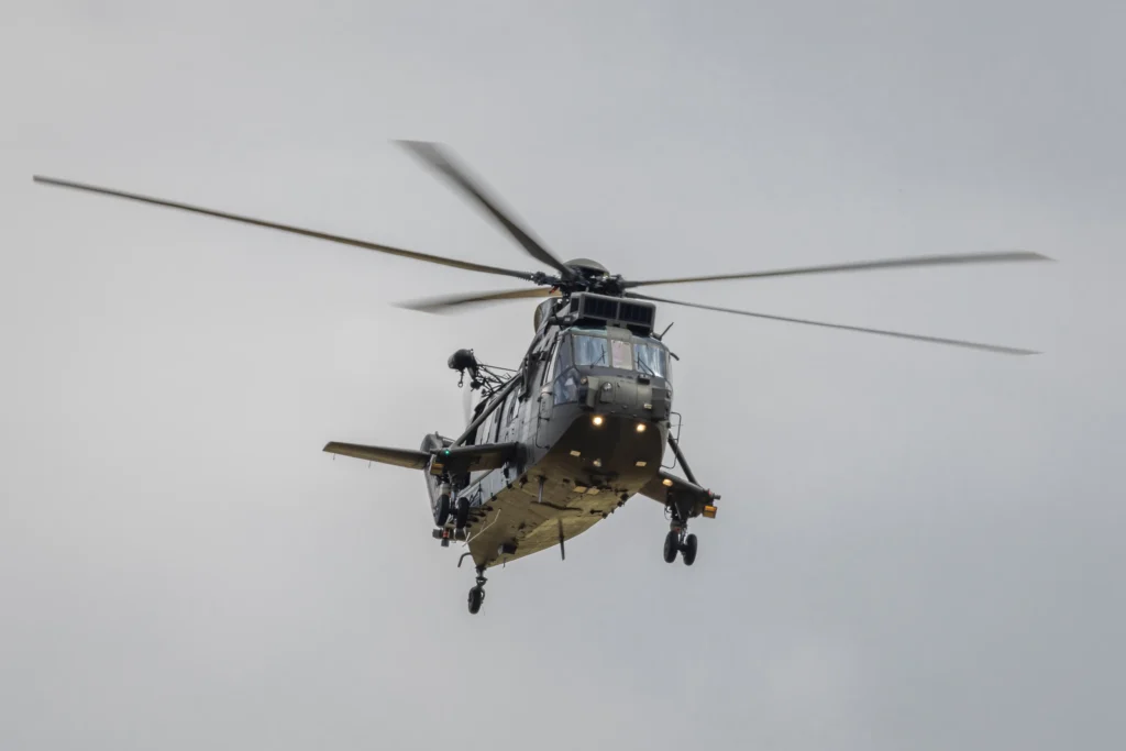 A military-style helicopter in flight against a grey, overcast sky. The helicopter's landing gear is down, lights are illuminated, and its rotors are captured mid-spin. The aircraft appears heavily equipped with external attachments and antennas.