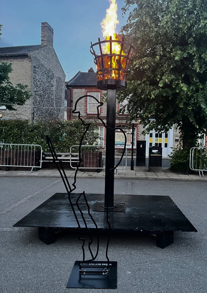 A lit beacon stands on a black platform in the middle of Thetford Marketplace at dusk. The flame burns brightly in a metal basket atop a tall post, with a silhouette sculpture of a soldier leaning on a rifle at the base. Surrounding buildings and trees frame the background, with metal barriers visible around the installation.