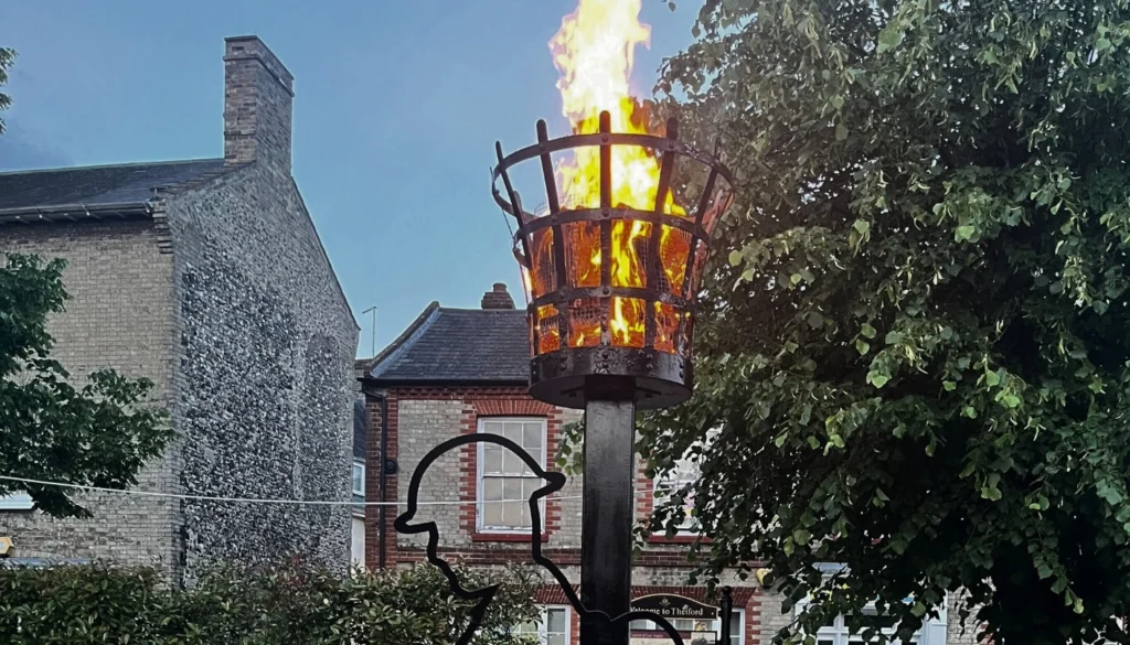A lit beacon stands on a black platform in the middle of Thetford Marketplace at dusk. The flame burns brightly in a metal basket atop a tall post, with a silhouette sculpture of a soldier leaning on a rifle at the base. Surrounding buildings and trees frame the background, with metal barriers visible around the installation.