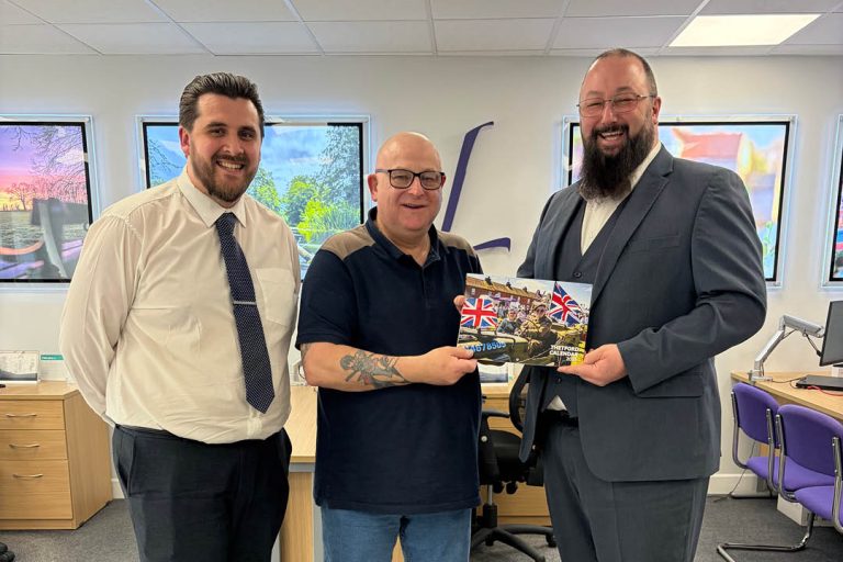 Three men stand in an office environment, smiling at the camera. The man in the centre holds a copy of the About Thetford calendar featuring a vintage jeep with Union Jack flags on the cover, while the men on either side stand close, dressed in smart-casual and formal attire. Desks, chairs and framed wall art are visible in the background.