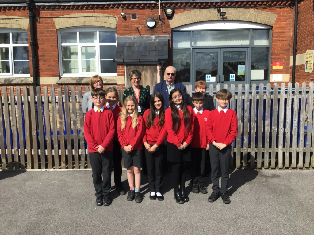 A group of primary school children and three adults stand outside Norwich Road Academy, posing for a group photo. The children, wearing red school jumpers and black trousers or skirts, smile in the sunlit schoolyard. Behind them is a wooden picket fence and a red-brick school building. Two women and a man (the Mayor and Mayoress of Thetford) stand behind the children, also smiling.