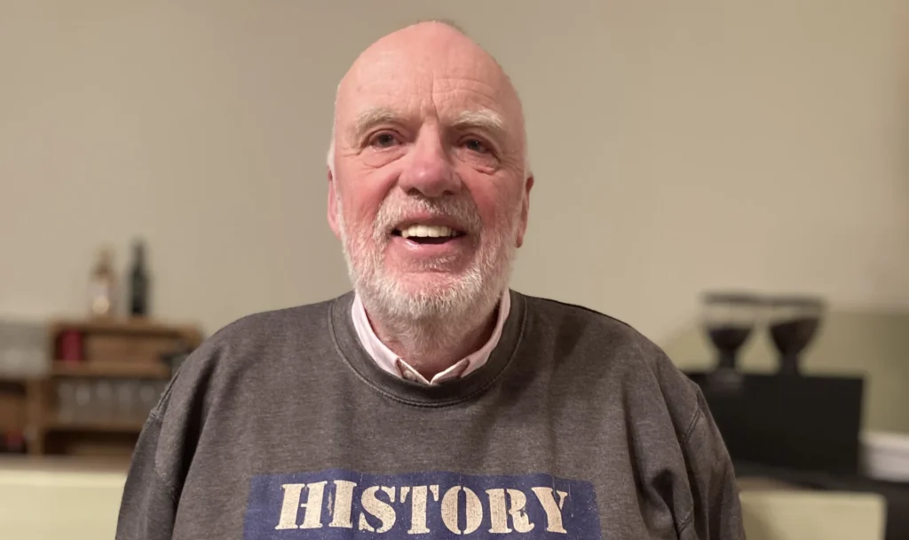 A smiling older man with a trimmed white beard and short hair, standing indoors against a pale wall. He is wearing a grey sweatshirt that features a distressed-style graphic across the chest reading "HISTORY MATTERS" — the word "HISTORY" is printed in white over a navy blue background, and "MATTERS" in white over a red background. His hands are tucked into his trouser pockets.