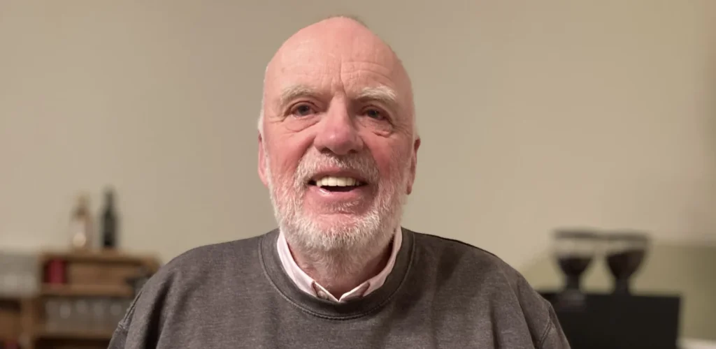 A smiling older man with a trimmed white beard and short hair, standing indoors against a pale wall. He is wearing a grey sweatshirt that features a distressed-style graphic across the chest reading "HISTORY MATTERS" — the word "HISTORY" is printed in white over a navy blue background, and "MATTERS" in white over a red background. His hands are tucked into his trouser pockets.