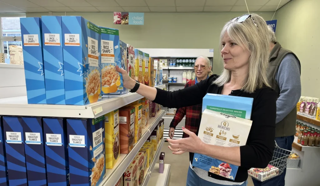 A woman with blonde hair, wearing a black top and blue jeans, stocks cereal boxes on a shelf in a small shop. She holds several other boxes against her chest. Behind her, two other shoppers, including an older man with a basket, browse the aisles. The shelves are stocked with various Tesco-branded cereal products.