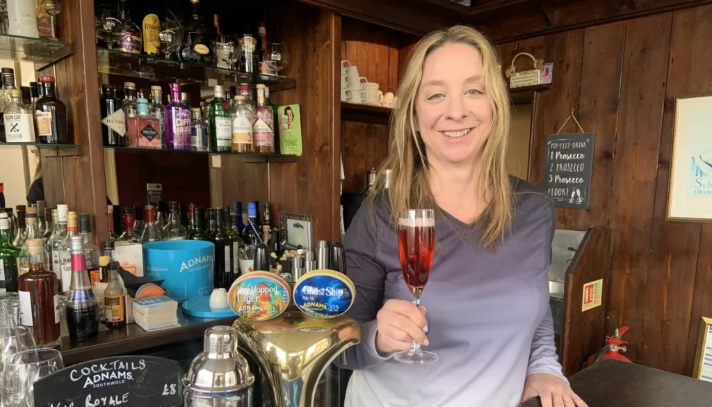A smiling woman stands behind a wooden bar holding a glass of pink cocktail. The bar is stocked with various spirits and mixers, including Adnams beer pumps and a cocktail shaker. A handwritten cocktail menu is visible on a small chalkboard.