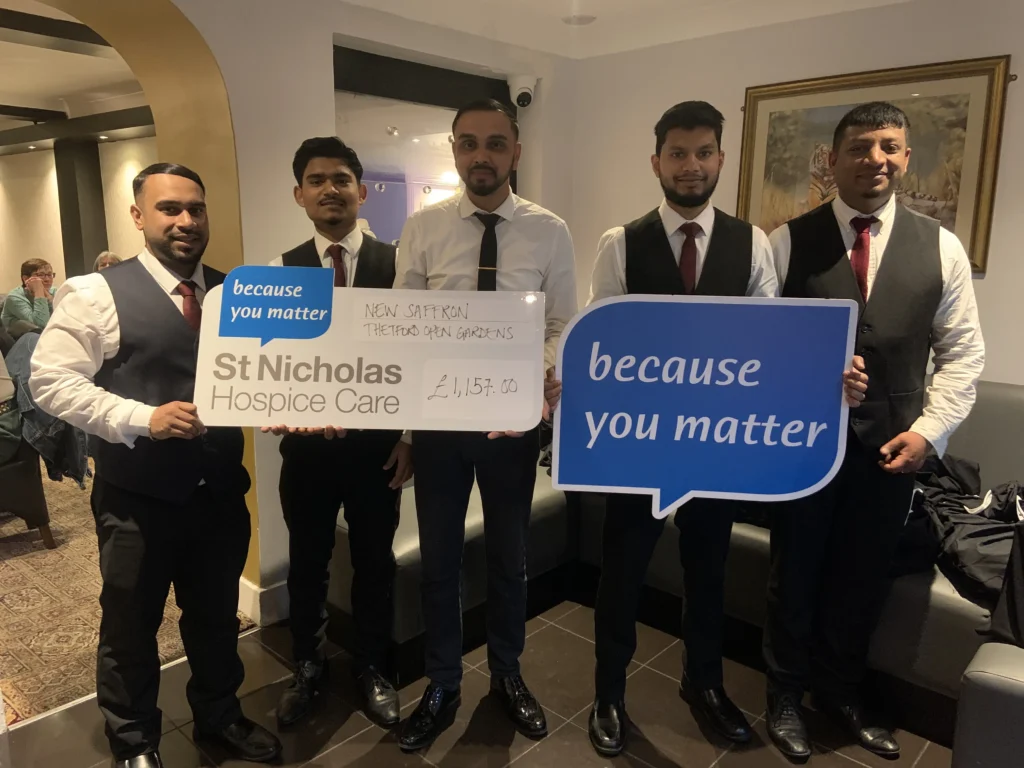 Five smartly dressed men from New Saffron restaurant stand together indoors, smiling and holding large signs reading "because you matter." They also display an oversized cheque for £1,157.00 made out to St Nicholas Hospice Care, raised through Thetford Open Gardens. Diners can be seen seated in the background.