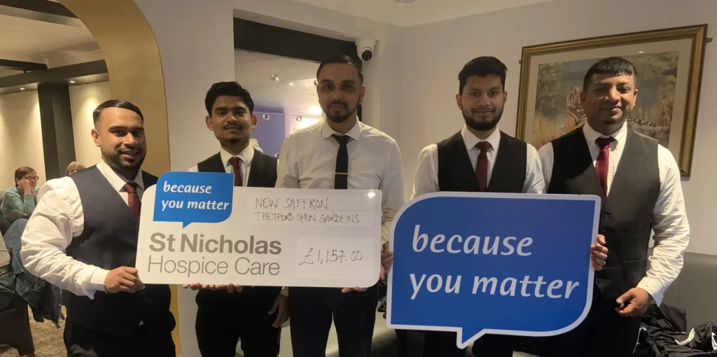 Five smartly dressed men from New Saffron restaurant stand together indoors, smiling and holding large signs reading "because you matter." They also display an oversized cheque for £1,157.00 made out to St Nicholas Hospice Care, raised through Thetford Open Gardens. Diners can be seen seated in the background.