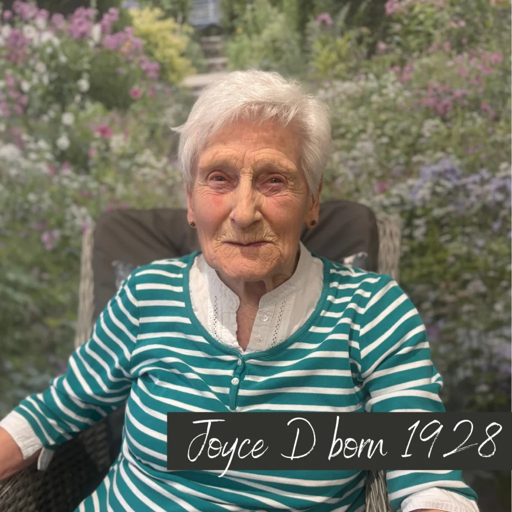 An elderly woman with white hair and a warm smile sits outdoors against a backdrop of a lush garden filled with purple and green foliage. She wears a green and white striped top with a white collar. Text on the image reads “Joyce D born 1928.”