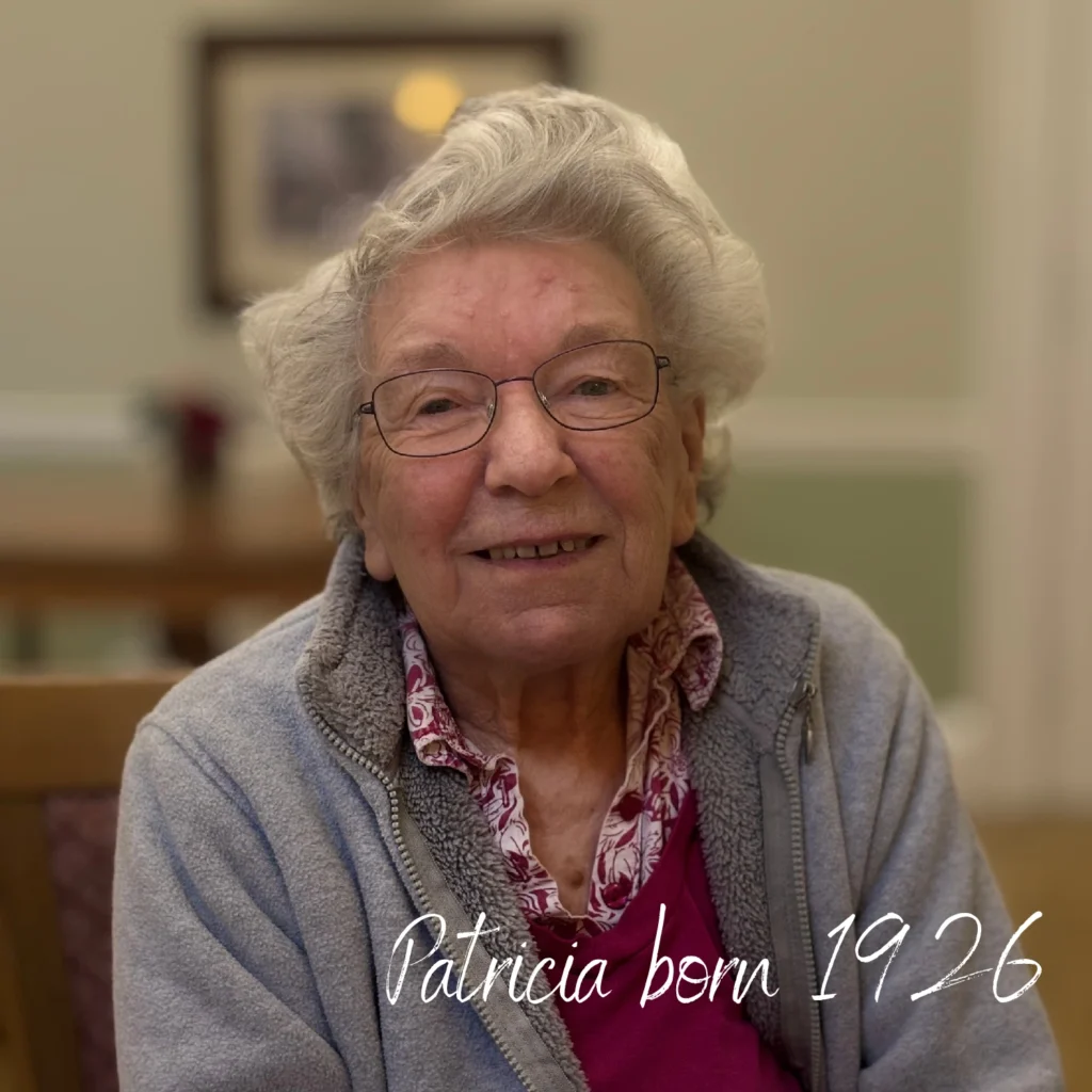 An elderly woman with short white hair and glasses smiles gently indoors, seated at a wooden table with a soft-focus background. She wears a light grey fleece over a red top with a floral shirt underneath. Text on the image reads “Patricia born 1926.”
