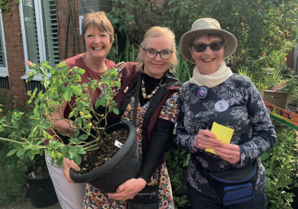 Three smiling women standing outdoors in a garden, holding a large potted plant. They are dressed casually and appear to be enjoying a community gardening or plant sale event. Behind them is a red-brick building with greenery and blooming plants.