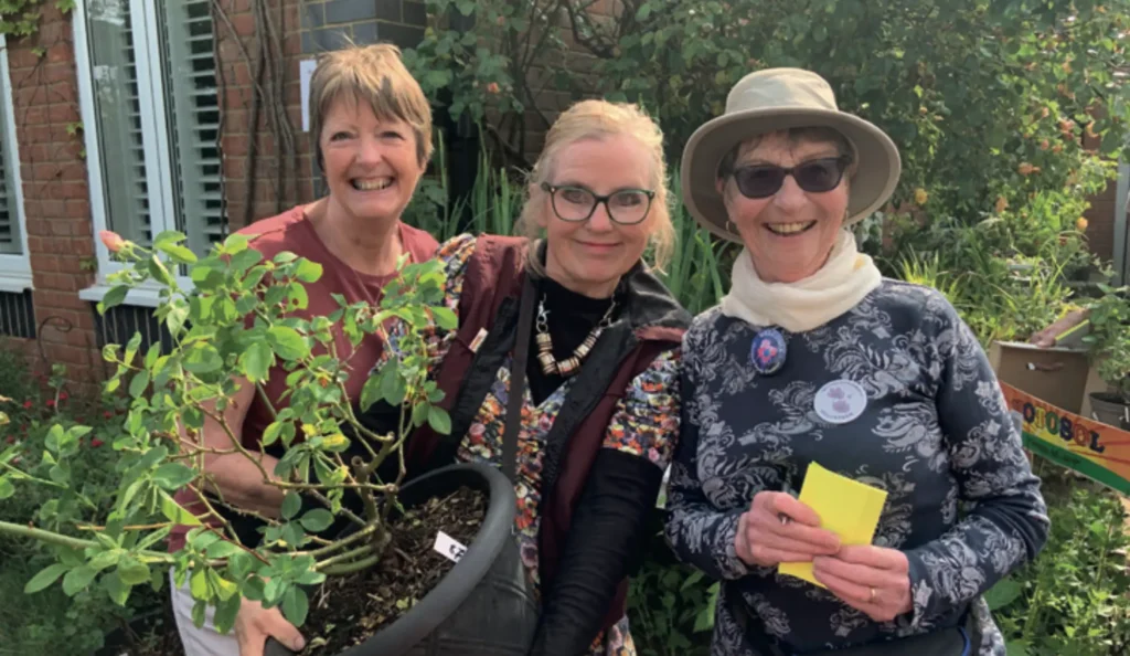 Three smiling women standing outdoors in a garden, holding a large potted plant. They are dressed casually and appear to be enjoying a community gardening or plant sale event. Behind them is a red-brick building with greenery and blooming plants.