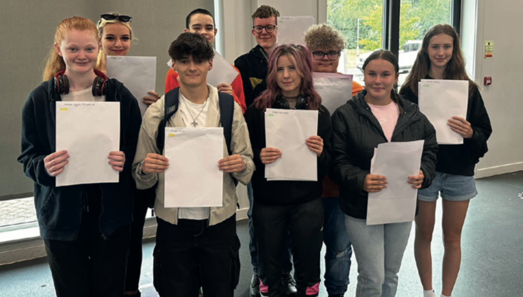A group of smiling students, standing indoors in two rows, proudly holding up white envelopes or certificates. They are dressed casually and appear to be celebrating exam results or achievements.