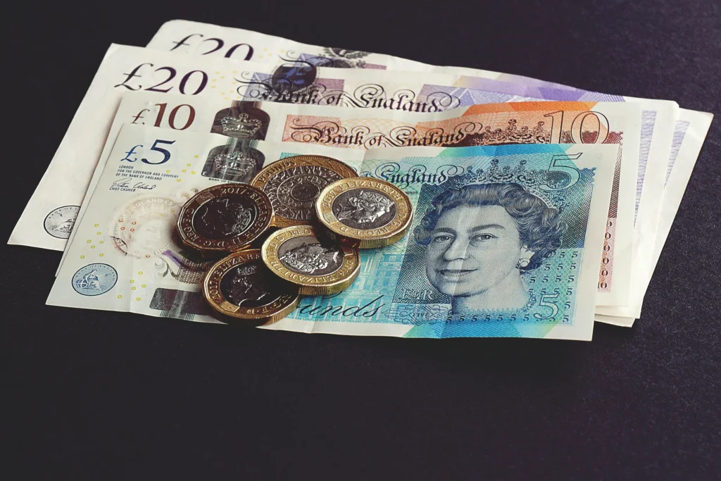 A close-up image of British currency featuring a £5 note with Queen Elizabeth II's portrait on top of a stack of other banknotes including £10 and £20 notes. Several £1 coins are scattered across the notes, all placed on a dark surface.