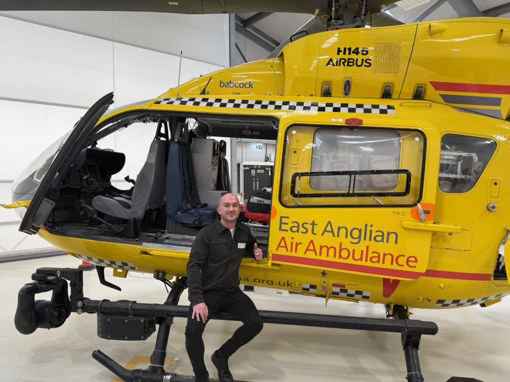 A man in a black uniform kneels beside a yellow East Anglian Air Ambulance helicopter inside a hangar. The helicopter door is open, revealing the cockpit and medical interior. The branding “East Anglian Air Ambulance,” “H145 Airbus,” and “babcock” are clearly visible on the side of the aircraft.