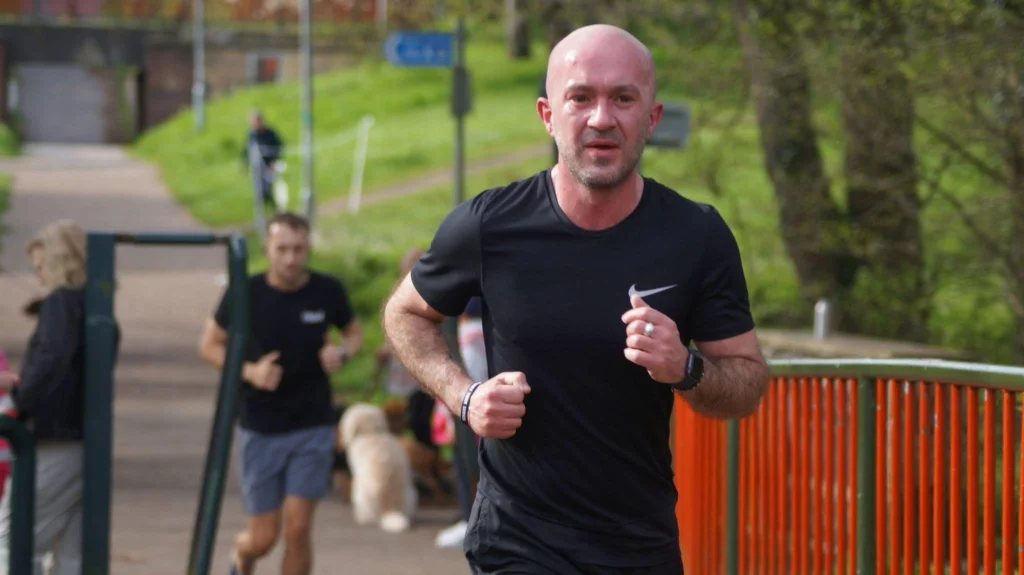 A man in a black Nike T-shirt runs towards the camera during what appears to be a park run event. He has a determined expression and is wearing a fitness watch and bracelet. Other runners and people, including someone with a dog, are visible in the blurred background, along with trees and a bright orange railing.