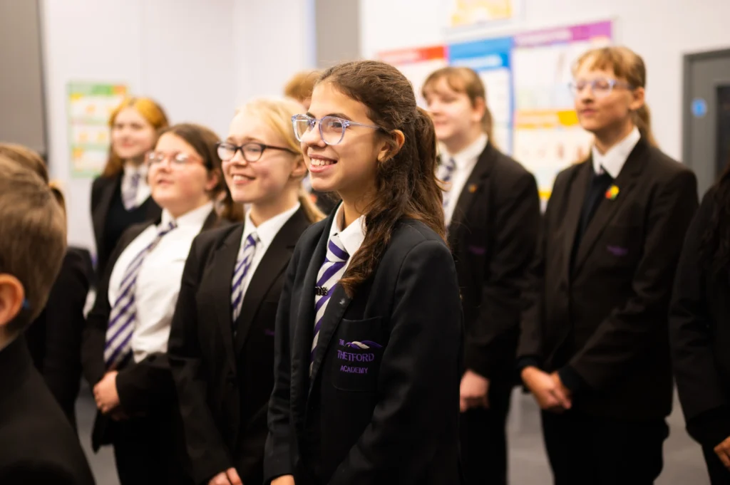 A group of smiling students in black uniforms and striped ties stand together in a classroom. The focus is on a girl in the front wearing glasses and a blazer embroidered with “The Thetford Academy.” Colourful educational posters are visible on the wall behind them.