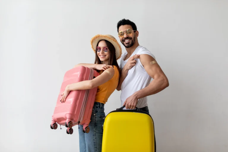 A smiling couple stands against a plain white background, each holding a brightly coloured suitcase—one pink, one yellow. They both wear sunglasses and casual holiday clothes, with the woman in a straw sunhat. Each person has a plaster on their upper arm, suggesting recent vaccinations. The man lifts his sleeve to show his plaster, and both appear ready for travel.