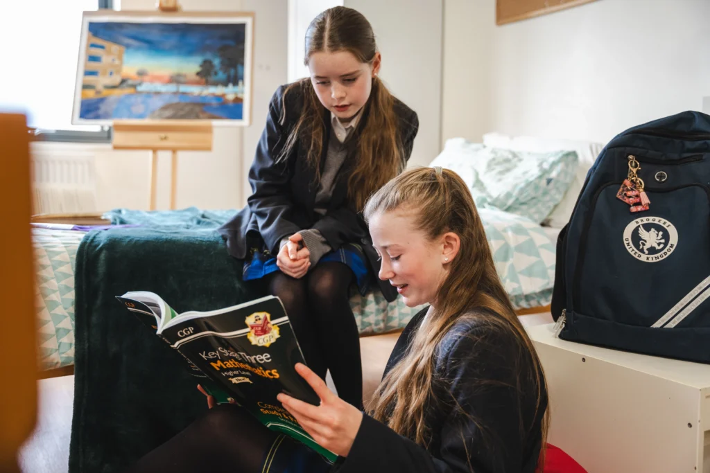 Two girls in school uniforms sit in a boarding room at Brookes UK. One reads from a CGP Maths revision book while the other looks on attentively. A Brookes UK school bag is placed on a white cabinet beside them, and an easel with colourful artwork is visible in the background.