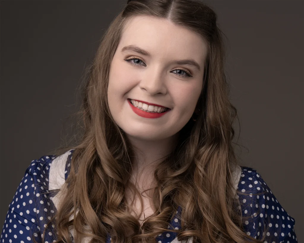Professional studio portrait of a smiling young woman with long, curled brown hair wearing a navy blue polka-dot dress with white trim. She has red lipstick, subtle eye makeup, and is posing against a plain grey background. The image has a soft, classic feel suited to vintage-themed promotion.