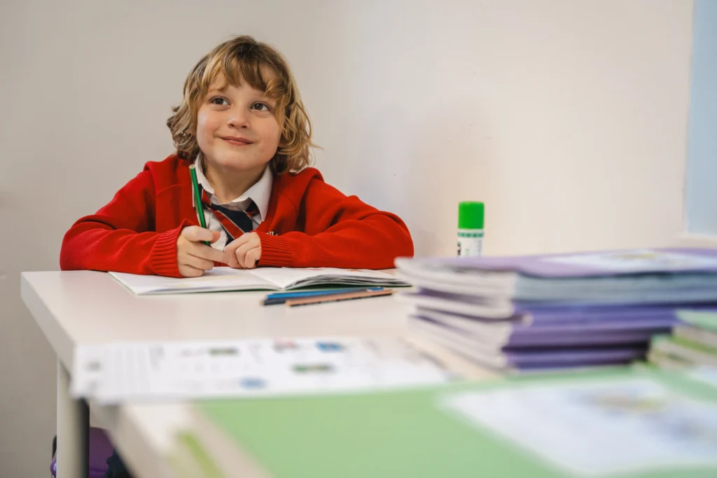 A young student in a red Brookes UK jumper and striped tie sits at a white desk, smiling while holding a green pencil. Exercise books are stacked in the foreground, and a glue stick and paper are also visible on the table.